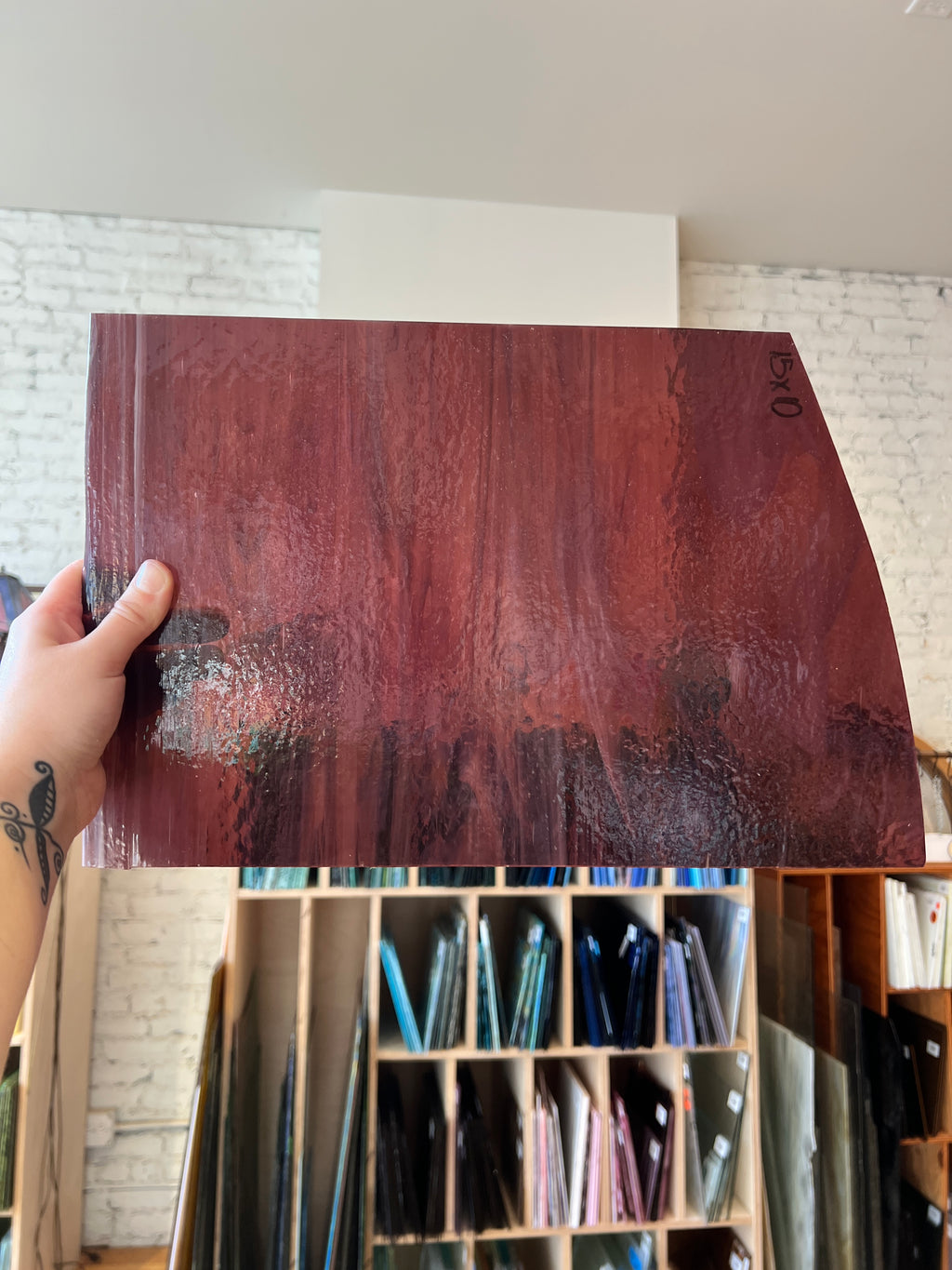 A hand holds up a large, glossy sheet of dark reddish-brown glass material, showing its reflective surface and texture. The background shows a workshop or storage room with shelves containing various colored material samples.