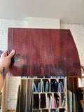 A hand holds up a large, glossy sheet of dark reddish-brown glass material, showing its reflective surface and texture. The background shows a workshop or storage room with shelves containing various colored material samples.