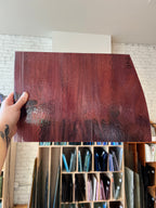 A hand holds up a large, glossy sheet of dark reddish-brown glass material, showing its reflective surface and texture. The background shows a workshop or storage room with shelves containing various colored material samples.