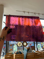 A hand holds a large rectangular stained glass panel with a textured, rippled design in shades of purple, magenta, and orange, set against a background of a stained glass studio with other colorful panels.