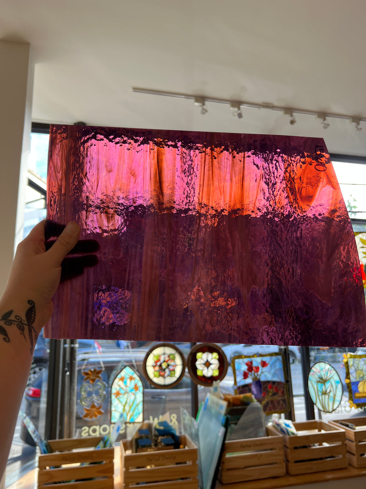 A hand holds a large rectangular stained glass panel with a textured, rippled design in shades of purple, magenta, and orange, set against a background of a stained glass studio with other colorful panels.
