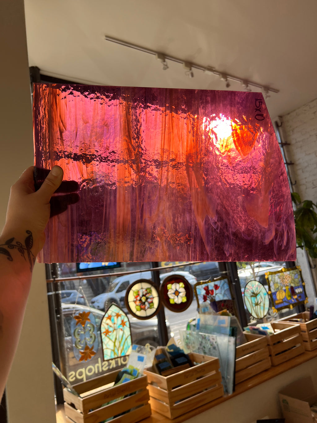 A hand holds up a large textured stained glass panel with a sunset design, showing vibrant pink, orange, and purple hues. In the background, a glass display case showcases other stained glass art pieces.