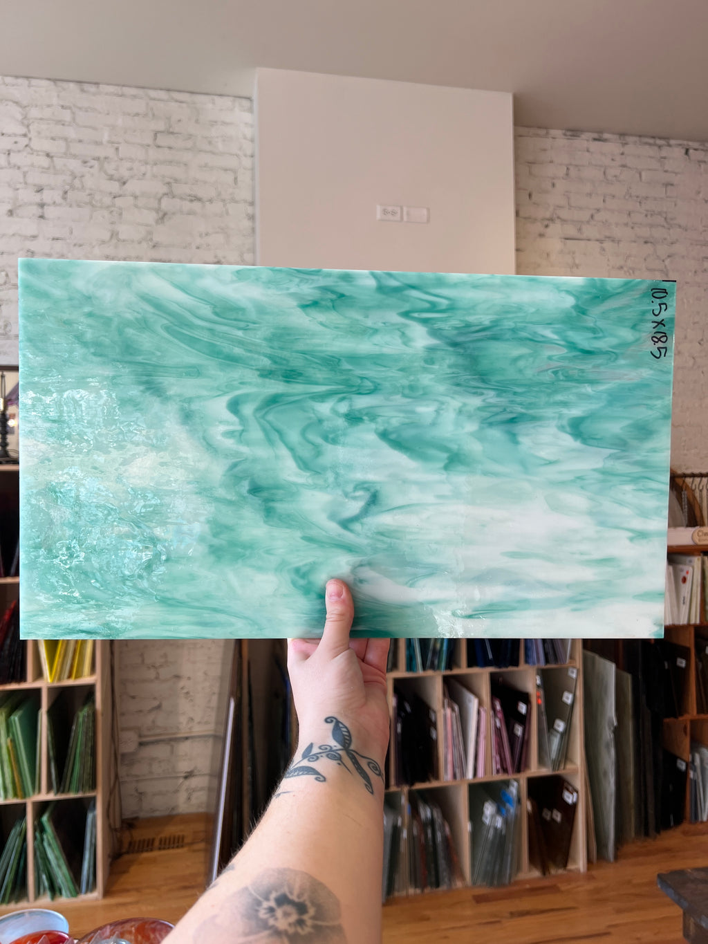 A hand holds up a large rectangular sheet of glass with a marbled teal, aqua, and white pattern, showing its 3x5 dimensions. The background is a workshop or studio with bookshelves and a white brick wall.