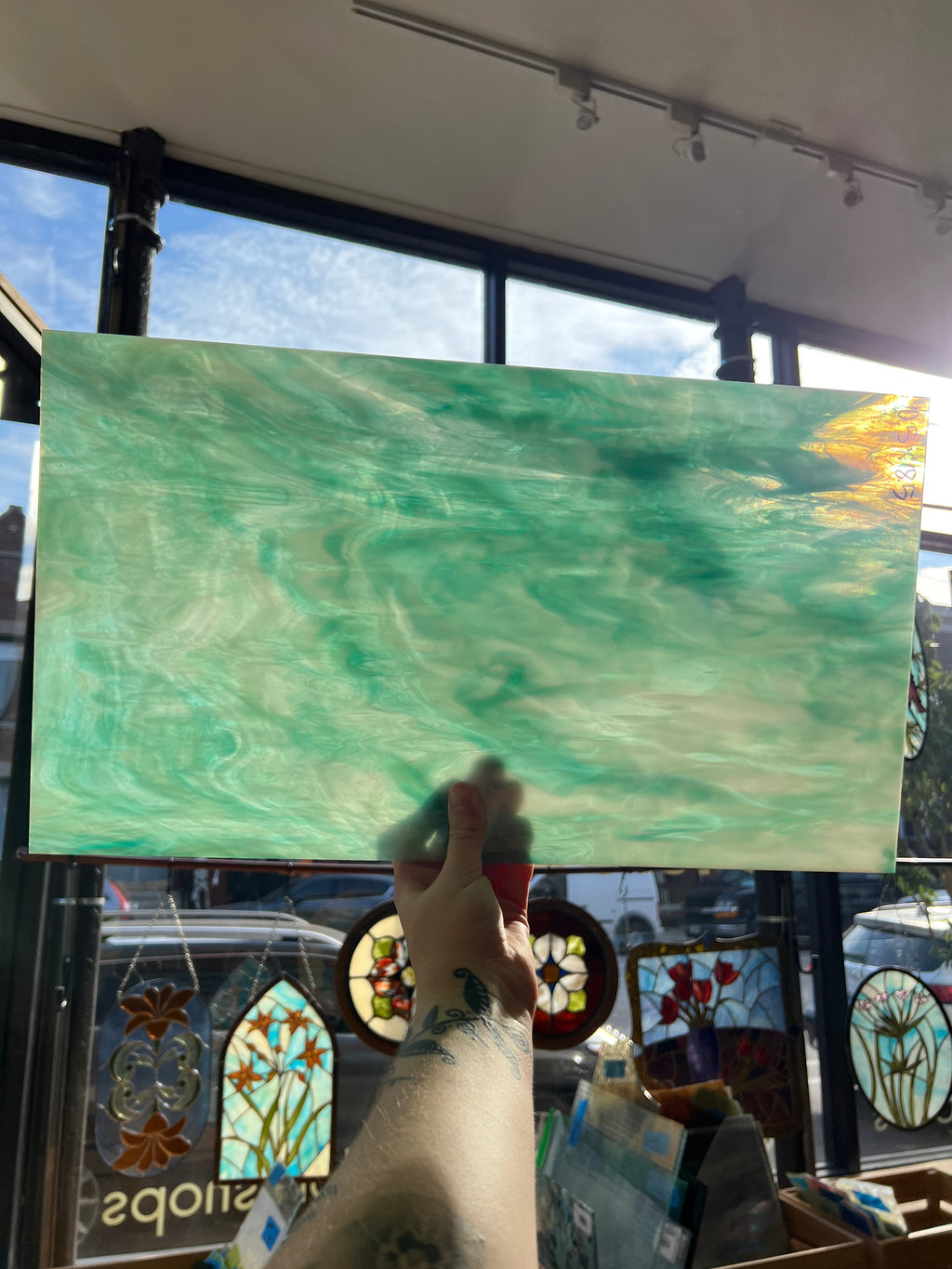 A hand holds a rectangular stained glass panel with a marbled green and turquoise pattern in front of a large window in a craft studio.