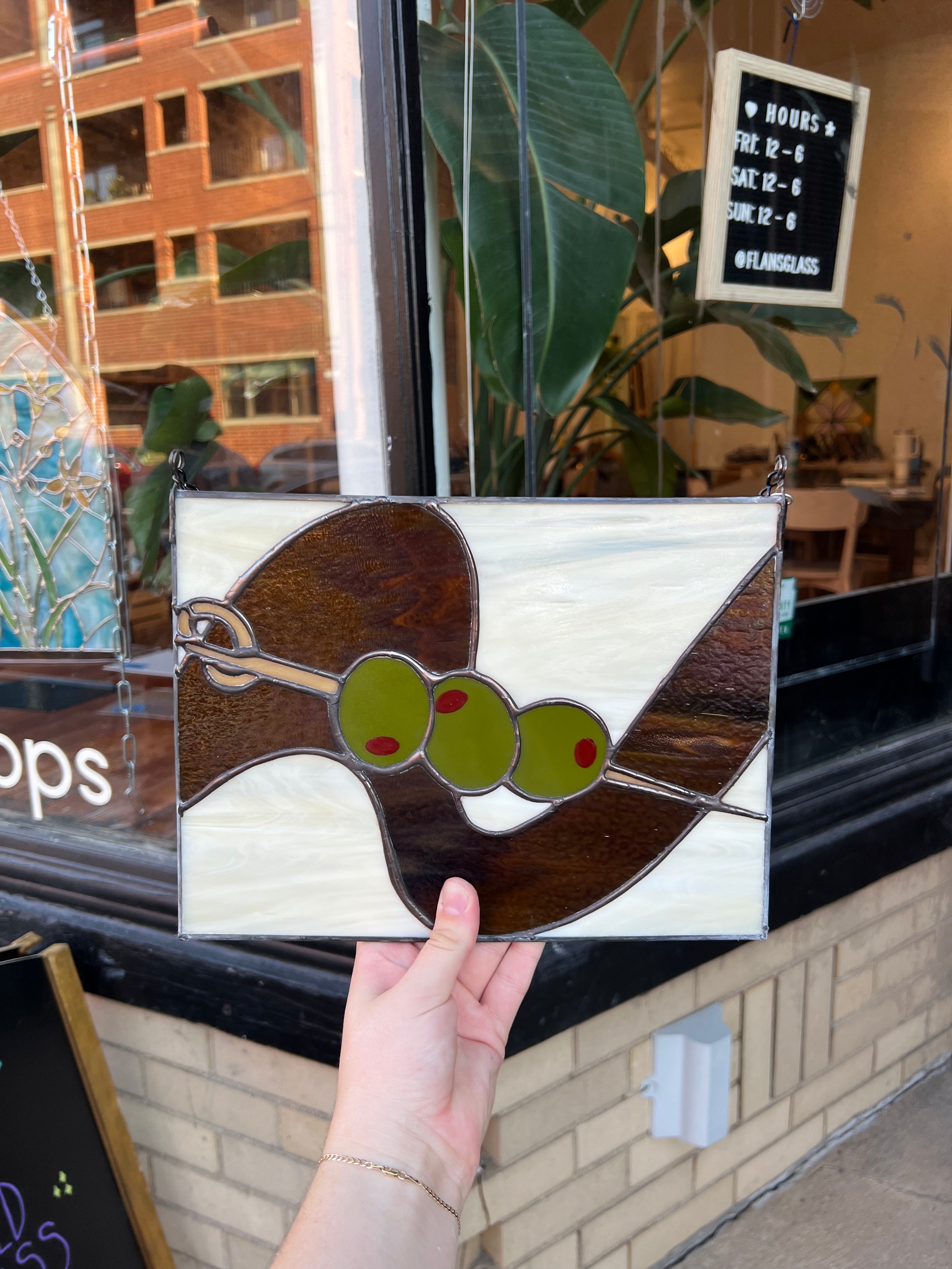 Hand holding a stained glass olive design in front of a store window.
