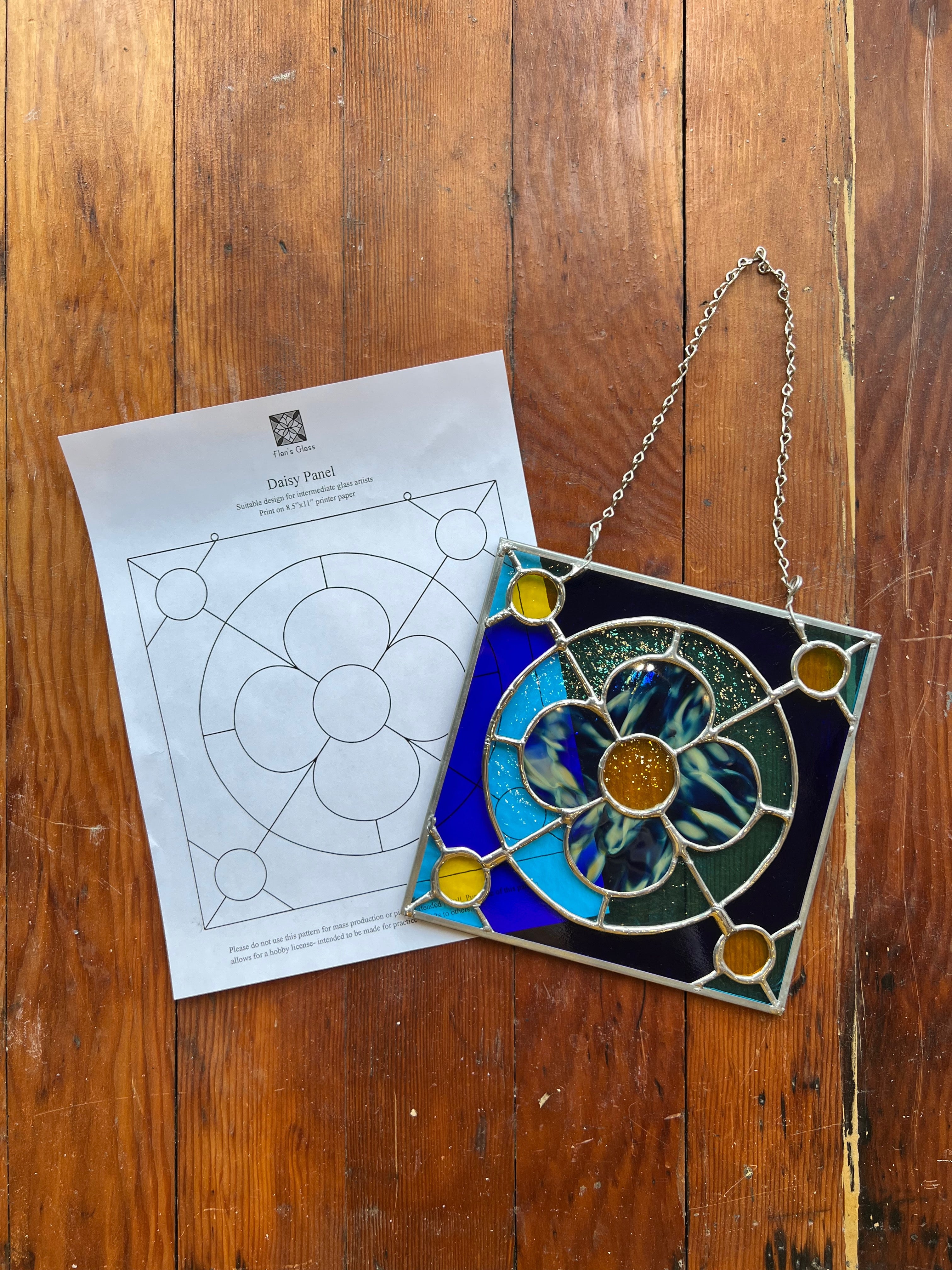 Stained glass panel with a chain on a wooden surface next to a pattern sheet.