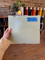 Hand holding a textured white piece of stained glass with a blue label in an art supply store.