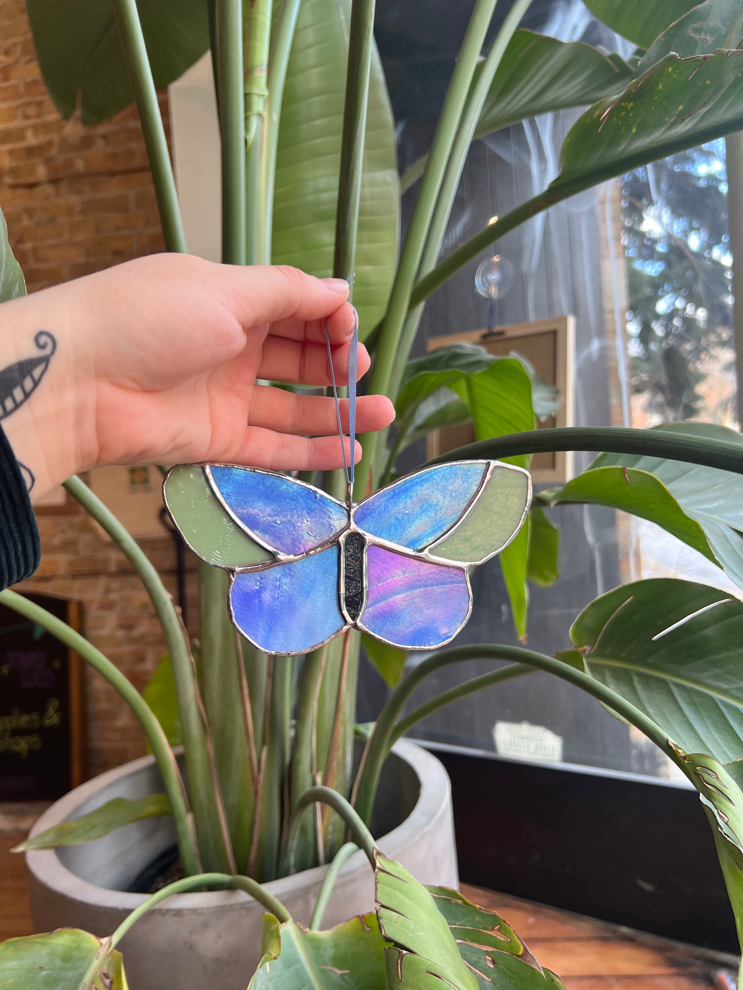 Hand holding a colorful butterfly-shaped stained glass art piece in front of a potted plant