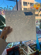 Hand holding a white sheet of mottled stained glass with a blue label in front of a colorful stained glass window.