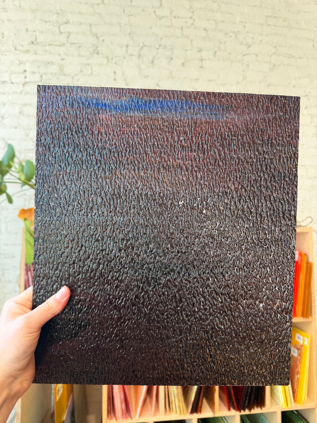 A hand holds a square black textured glass sample, showing a pebbled or grainy surface, against a light brick wall background with shelves of colorful items below.