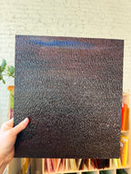 A hand holds a square black textured glass sample, showing a pebbled or grainy surface, against a light brick wall background with shelves of colorful items below.