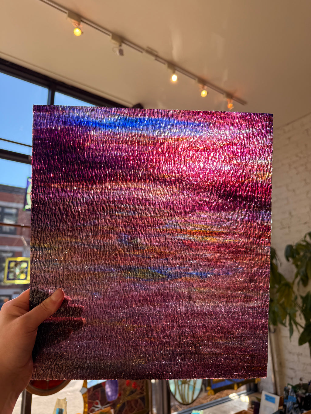 A hand holds a square textured piece of glass featuring an abstract, shimmering sunset-like pattern with pink, purple, blue, and orange tones, photographed indoors with a window and track lighting in the background.