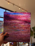 A hand holds a square textured piece of glass featuring an abstract, shimmering sunset-like pattern with pink, purple, blue, and orange tones, photographed indoors with a window and track lighting in the background.