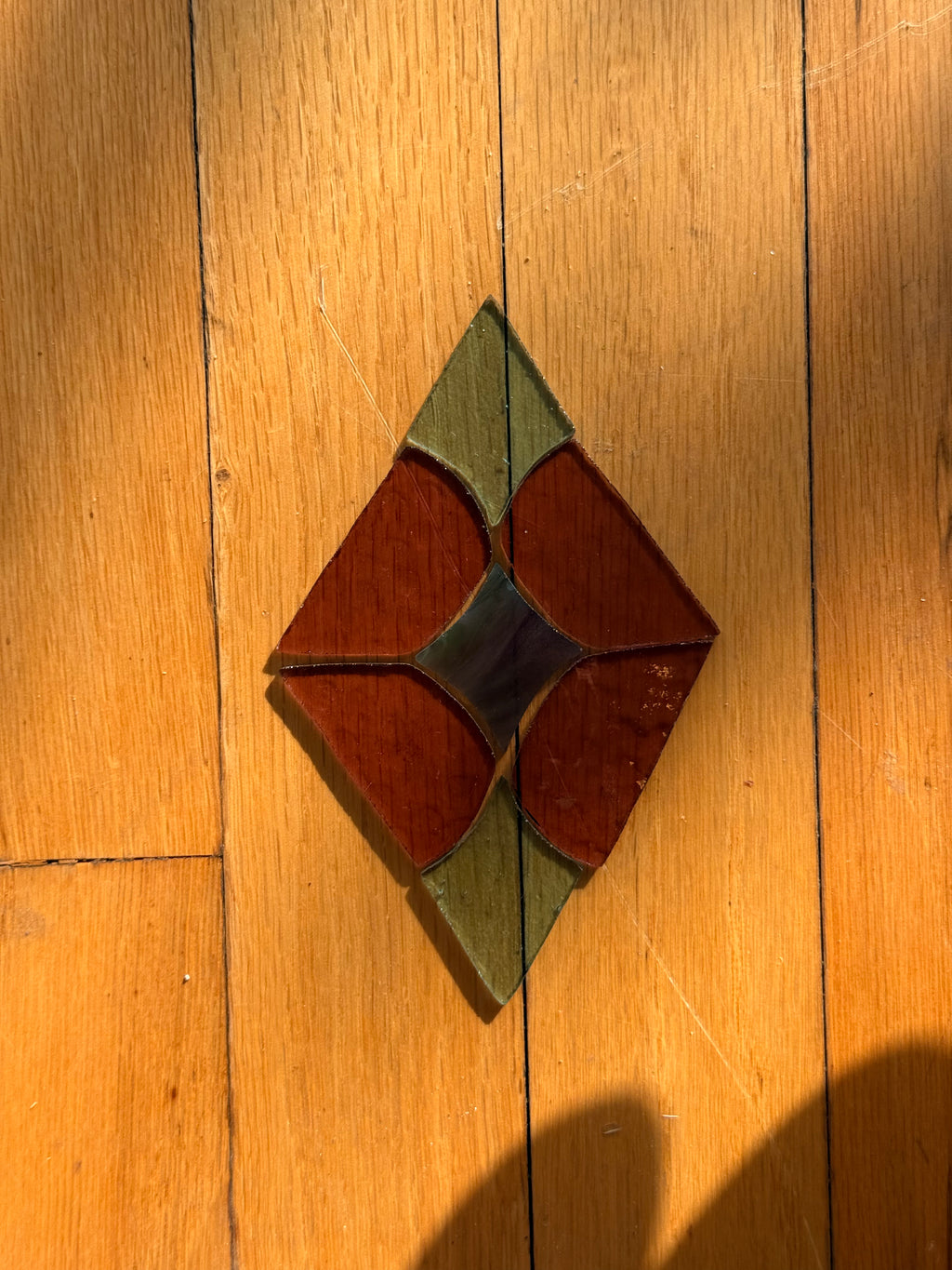 A diamond-shaped stained glass pre cut kit with a diamond pattern, resting on a wooden plank floor under warm sunlight.