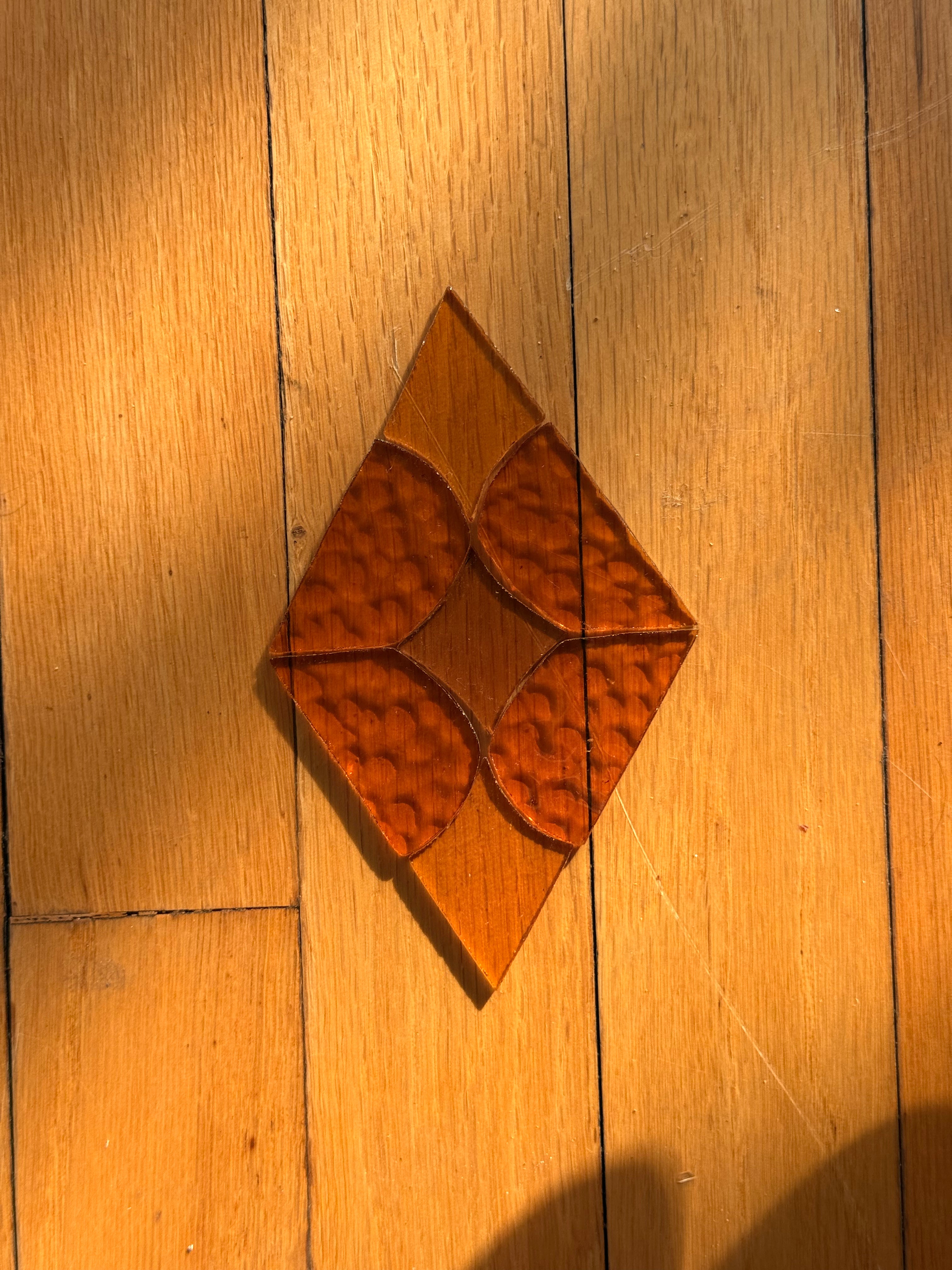 A diamond-shaped stained glass pre cut kit with a diamond pattern, resting on a wooden plank floor under warm sunlight.