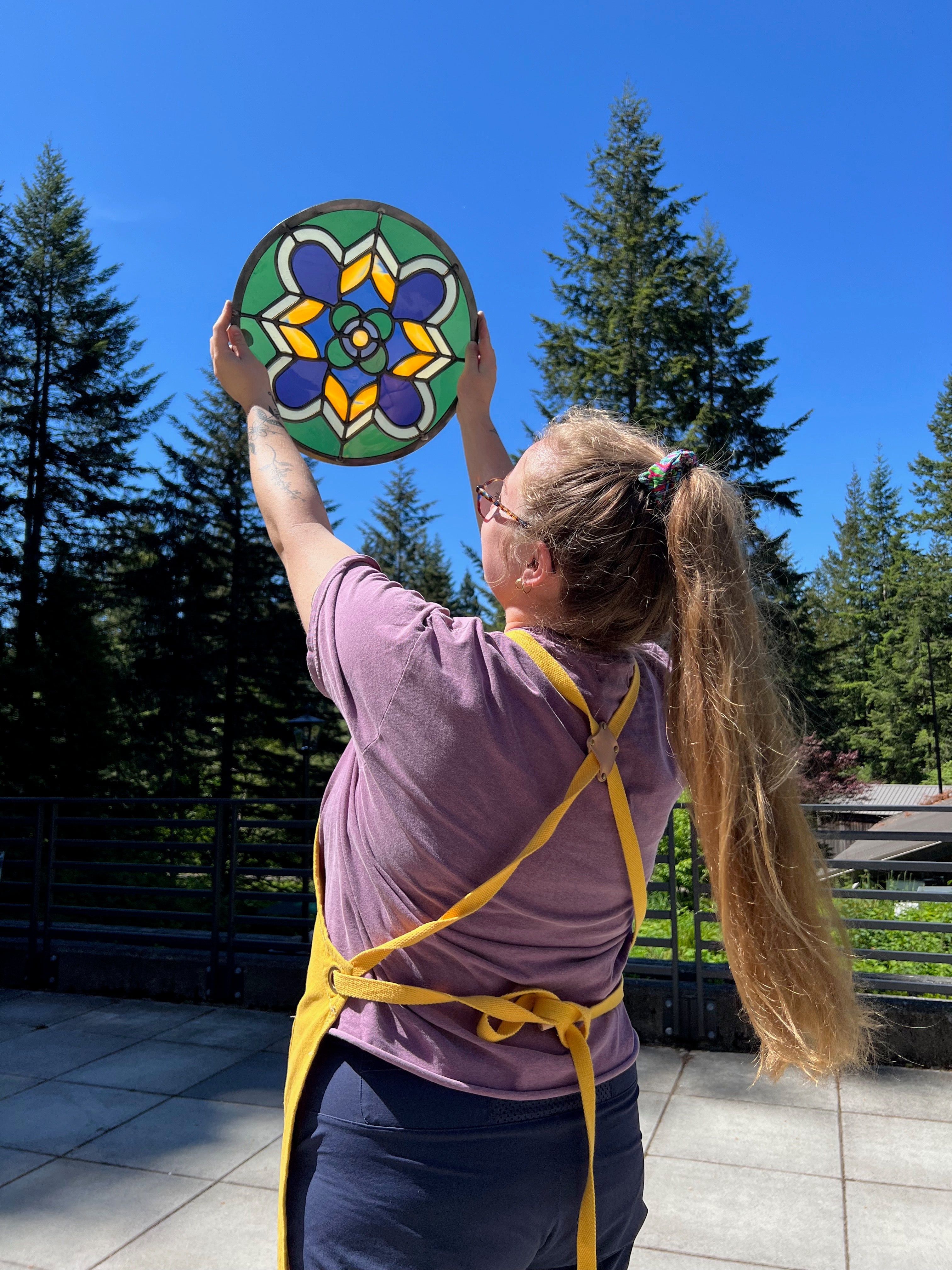 Person holding a colorful stained glass mandala against a blue sky with trees in the background