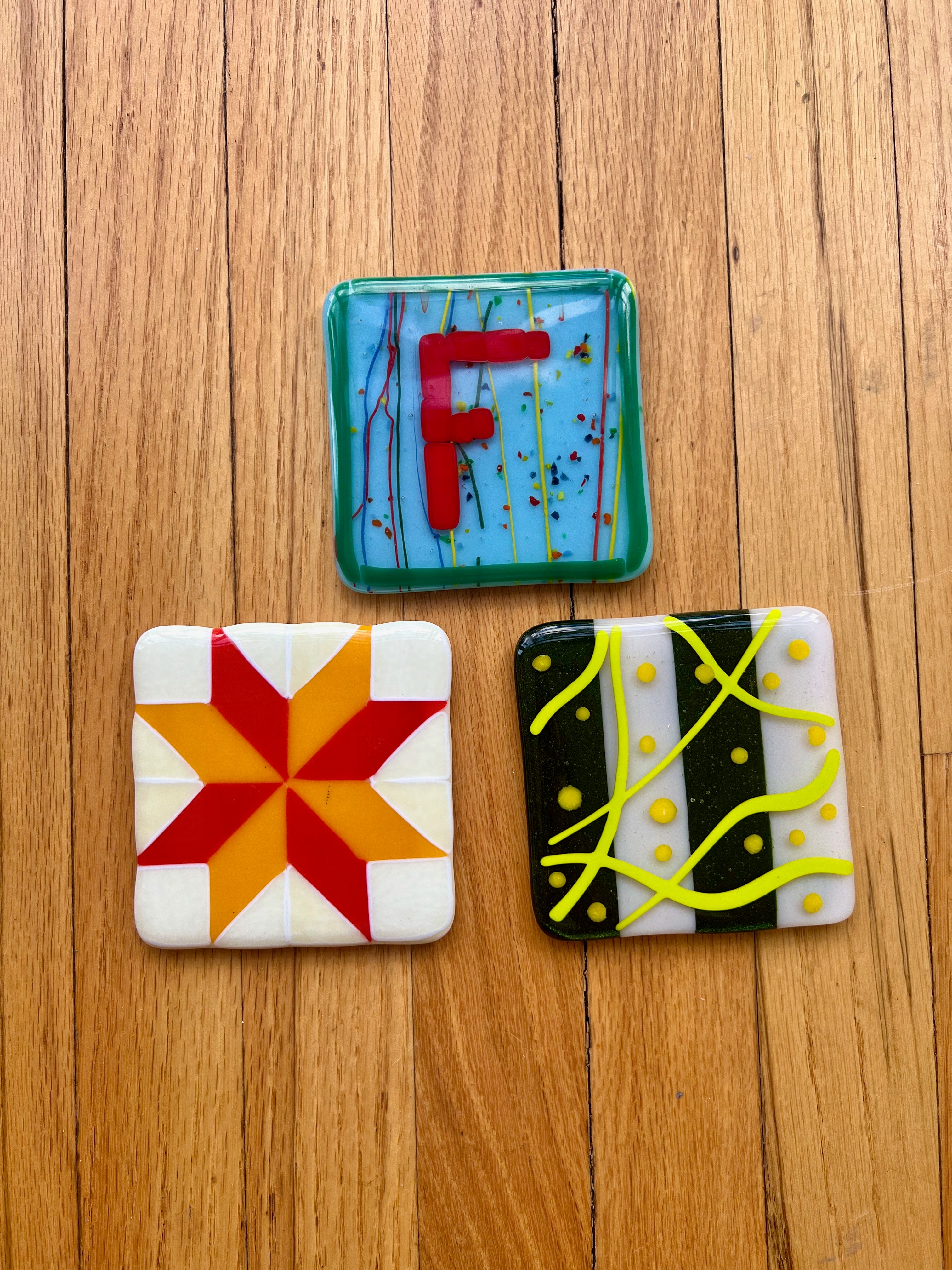 Three colorful square fused glass coasters on a wooden surface