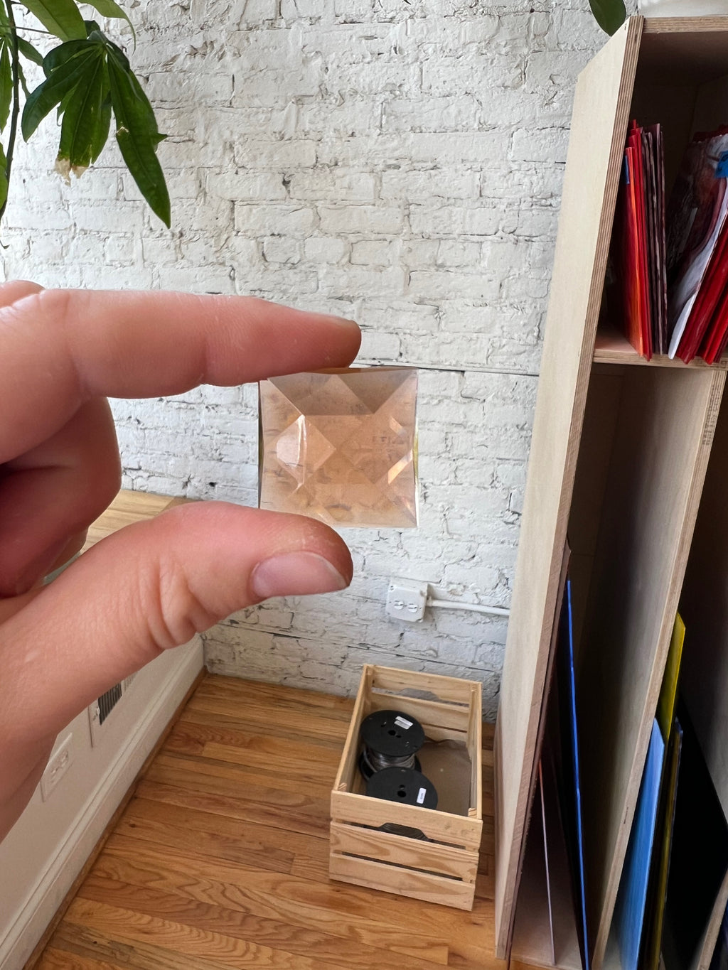 Hand holding a small transparent gem with a brick-patterned wall and wooden shelf in the background.