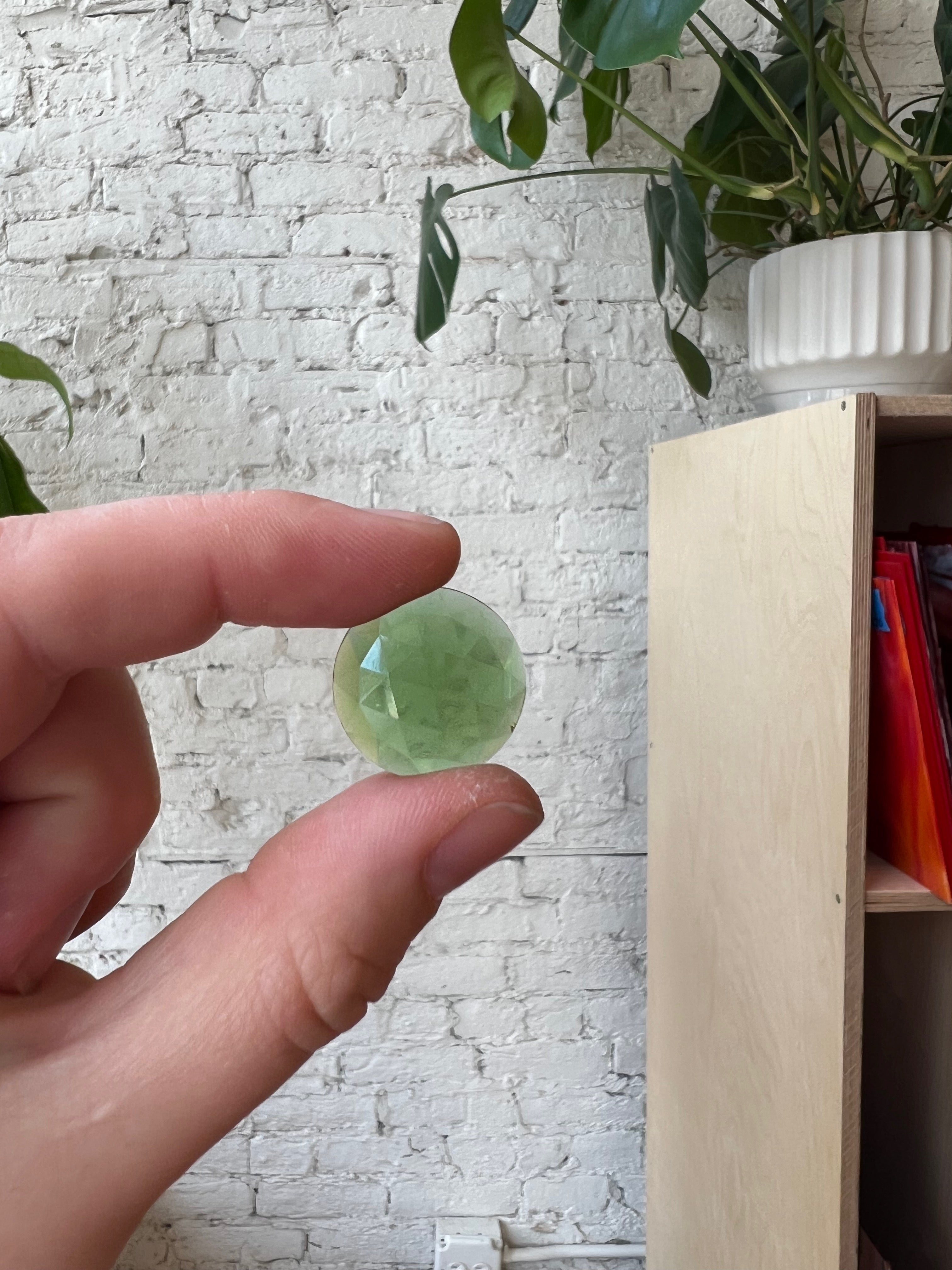 Hand holding a green gemstone in front of a white brick wall and wooden shelf.