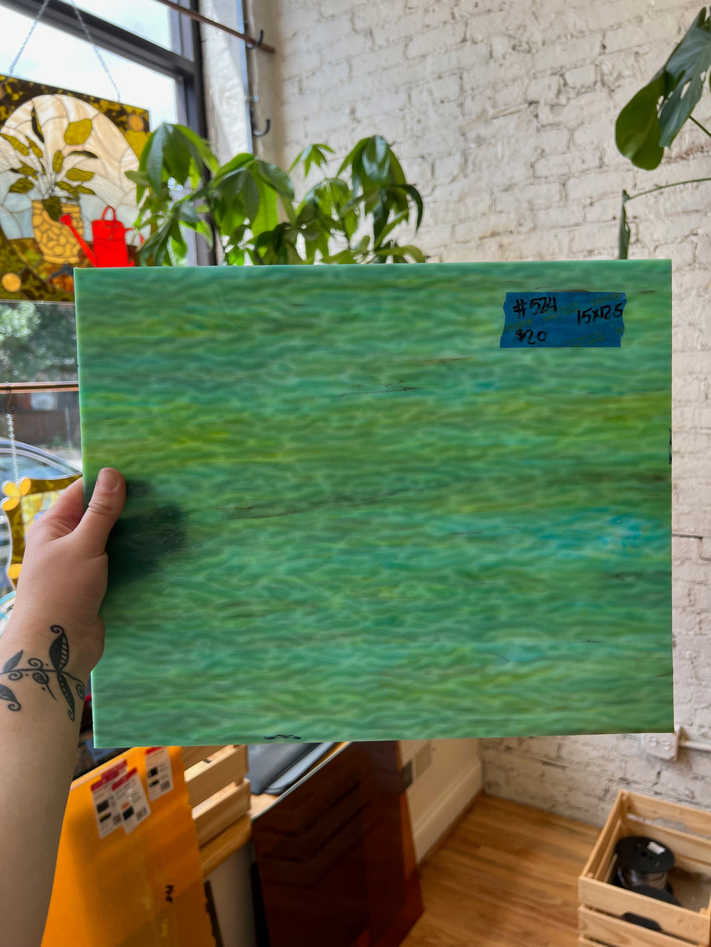 Person holding a green textured piece of glass indoors with plants and a window in the background.