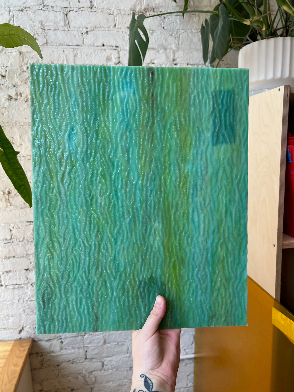 Hand holding a green textured glass against a white brick wall with plants.