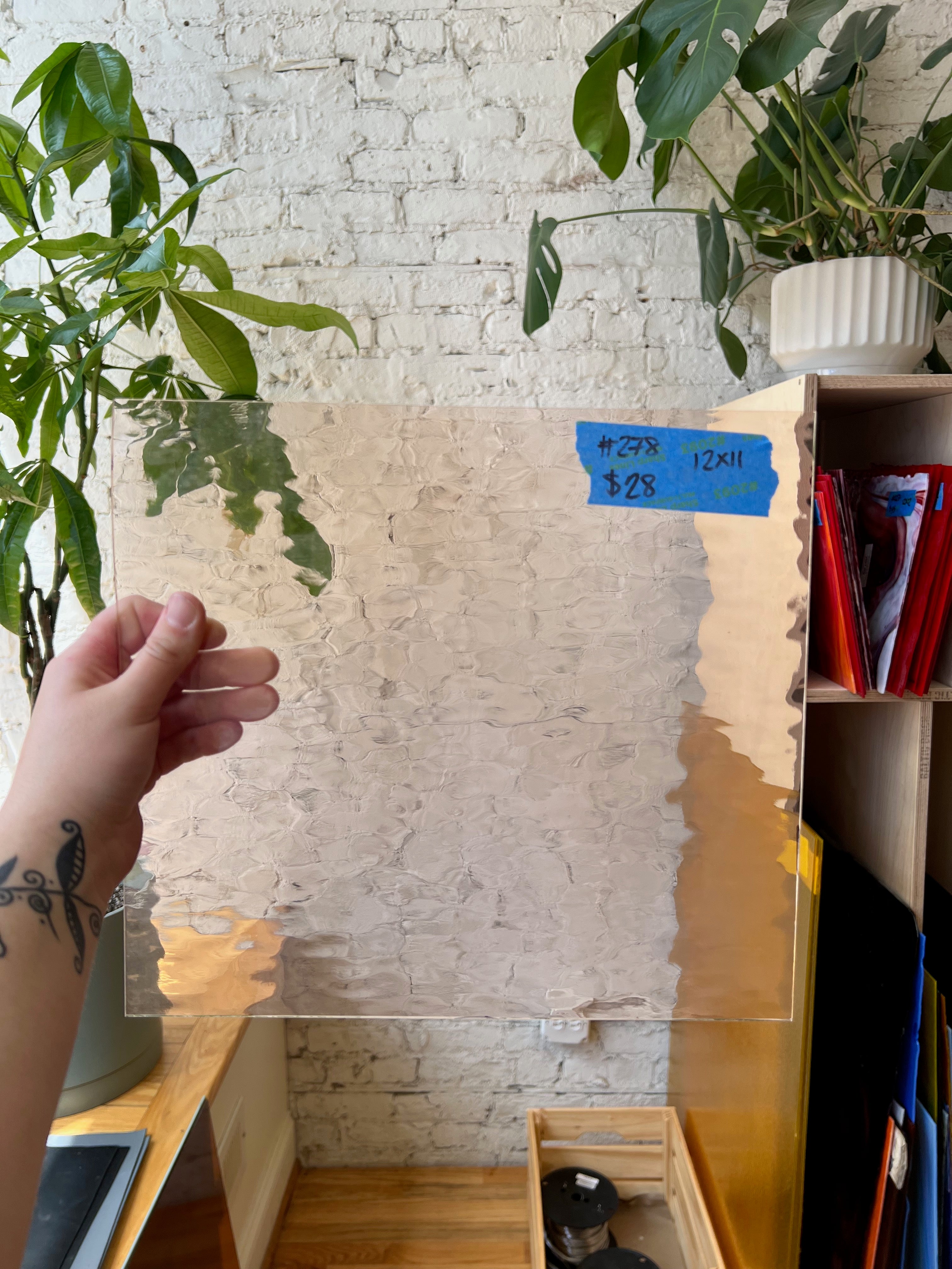 Clear textured glass sheet held by a hand with a white brick wall and plants in the background