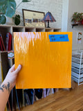 Orange sheet of glass held by a hand in a room with a lamp and books.
