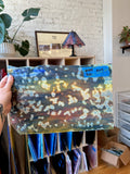 Hand holding a colorful abstract glass sheet in a room with shelves and plants.