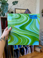 Hand holding a glass sheet with green and blue wavy patterns in an indoor setting.
