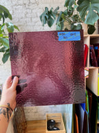 Person holding a textured purple glass sheet with a brick wall and plants in the background