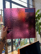 Hand holding a textured pink and purple glass sheet in front of a window with stained glass