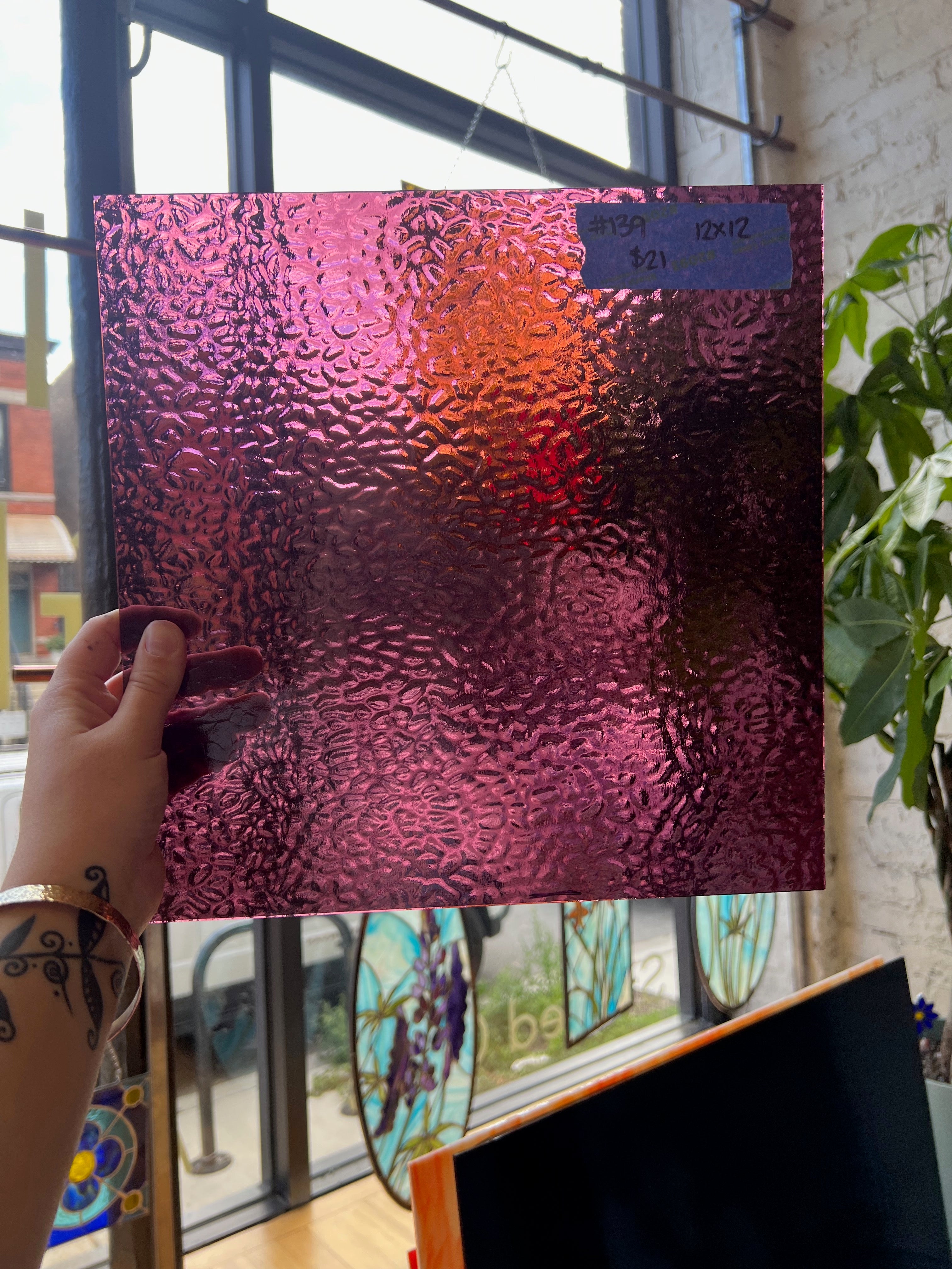 Hand holding a textured pink and purple glass sheet in front of a window with stained glass