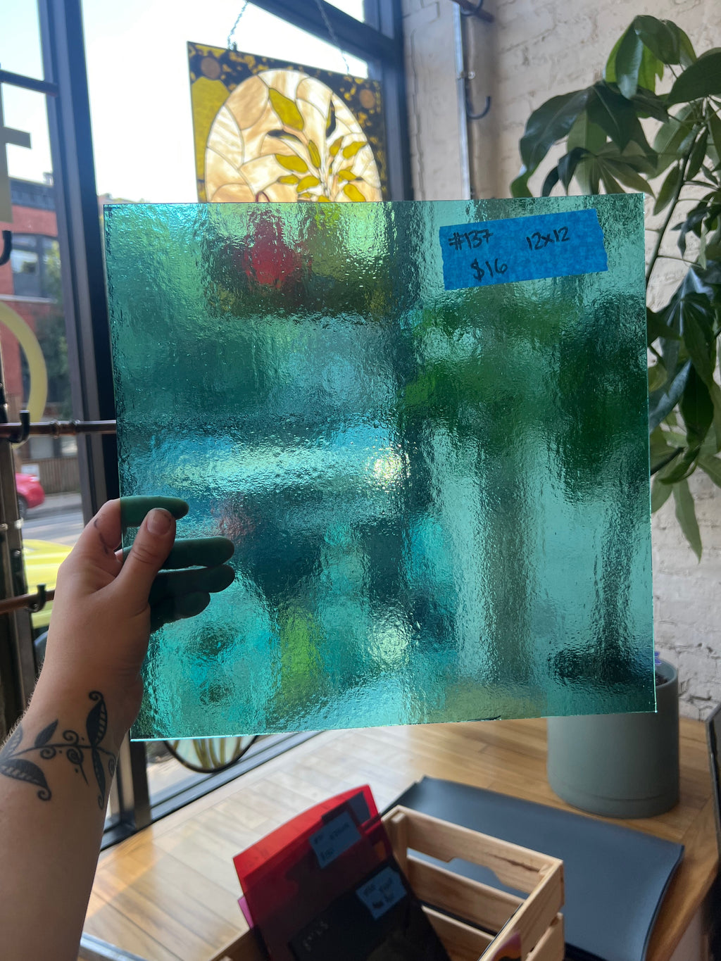 Turquoise glass panel held by a hand with a tattoo, in front of a window with stained glass and plants.