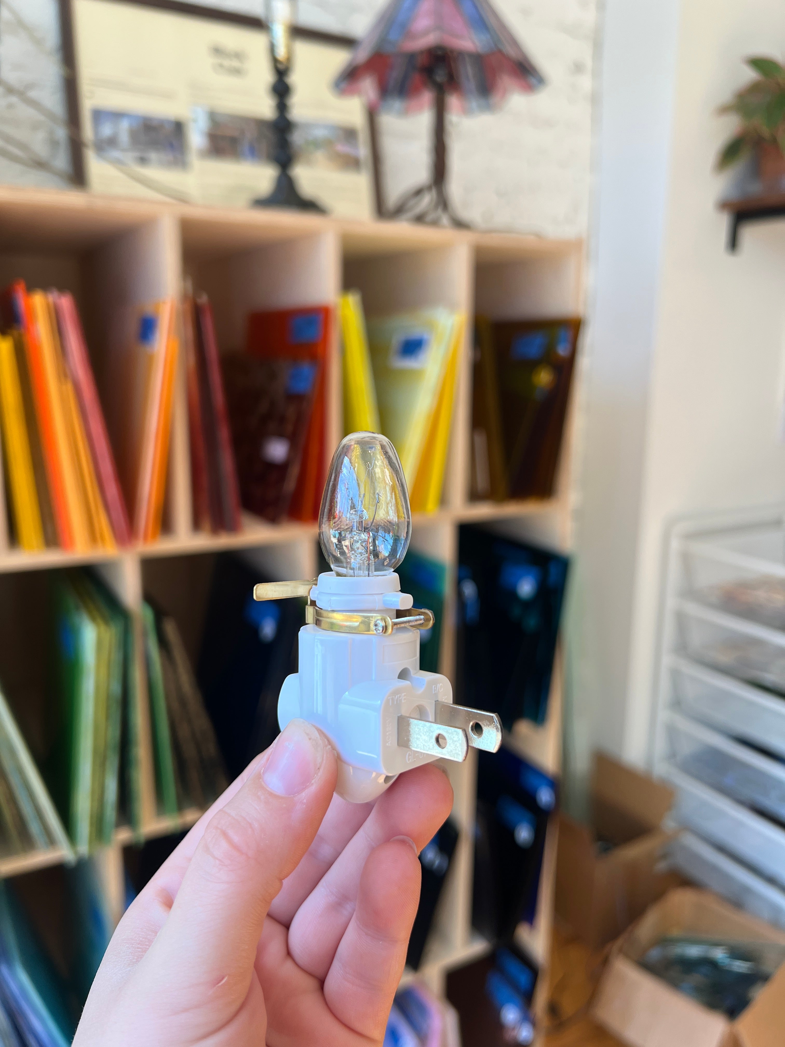 Hand holding a light bulb and plug against a blurred background of books and stationery items.