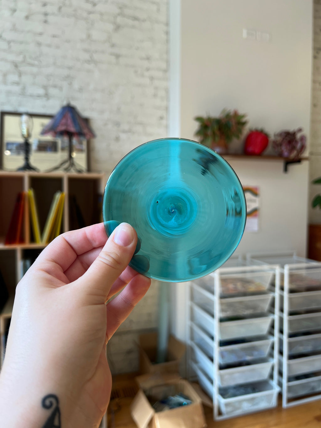 Hand holding a teal glass rondel in an indoor setting with shelves and boxes in the background.