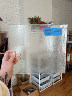 Person holding a square piece of stained glass with a blue label in an indoor setting.