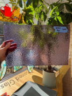 Person holding a sheet of textured lilac glass with a plant and window in the background