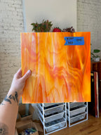Hand holding an orange swirled piece of glass against a brick wall background. 