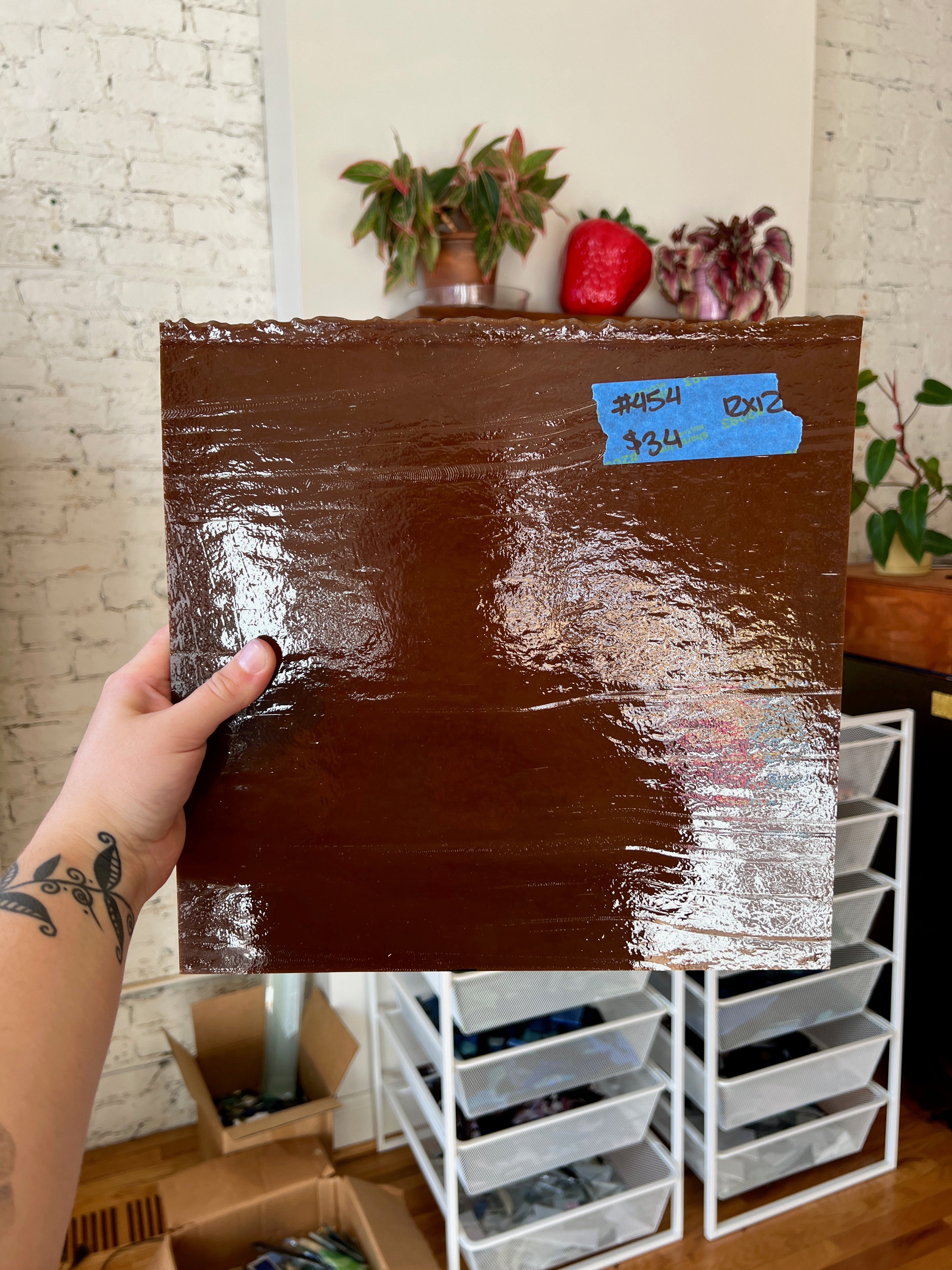 Person holding a large brown sheet of glass with a white brick wall and storage shelves in the background.