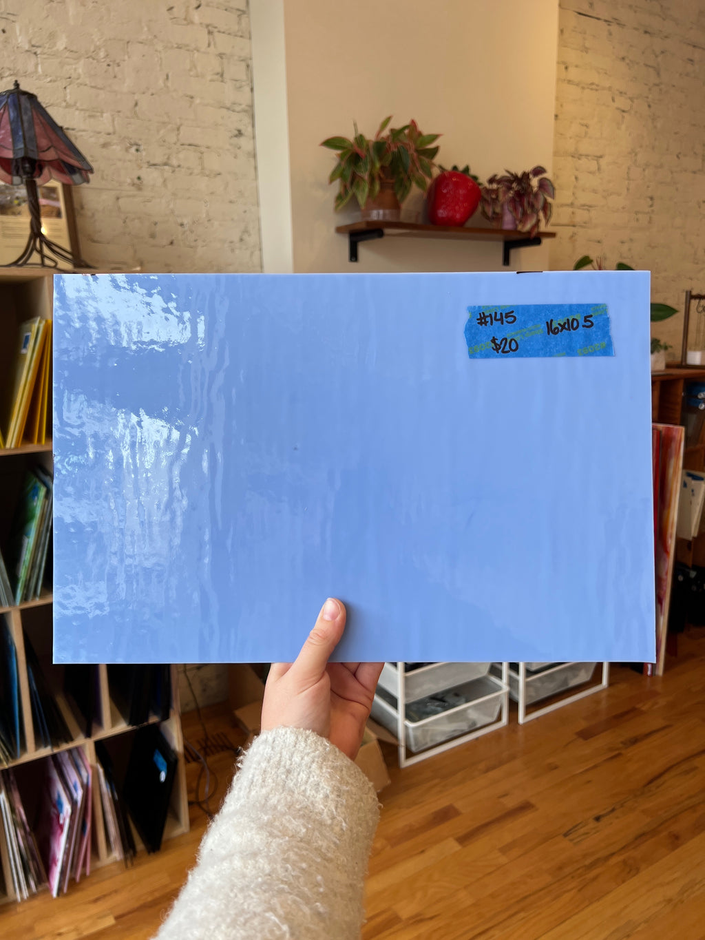 Hand holding a blue piece of stained glass in a room with a bookshelf and plants.