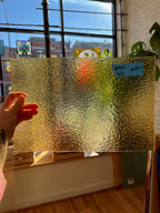 Gold textured glass sheet held by a hand with a window and plant in the background
