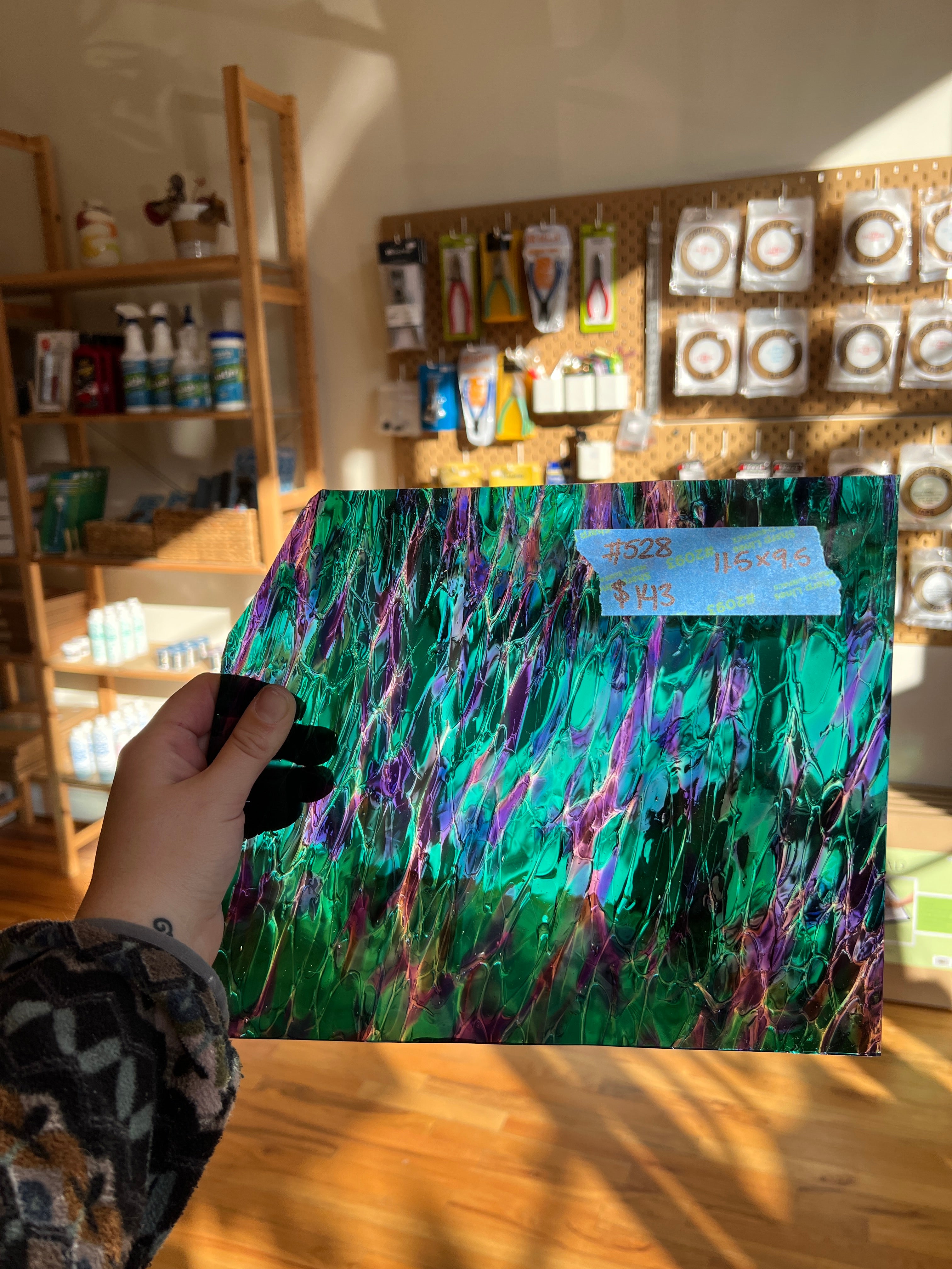 Hand holding a colorful abstract glass sheet in front of a display of art supplies.