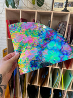 Hand holding a colorful, iridescent sheet of glass in front of a bookshelf.