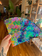 Hand holding a colorful abstract glass sheet in a room with shelves and plants.