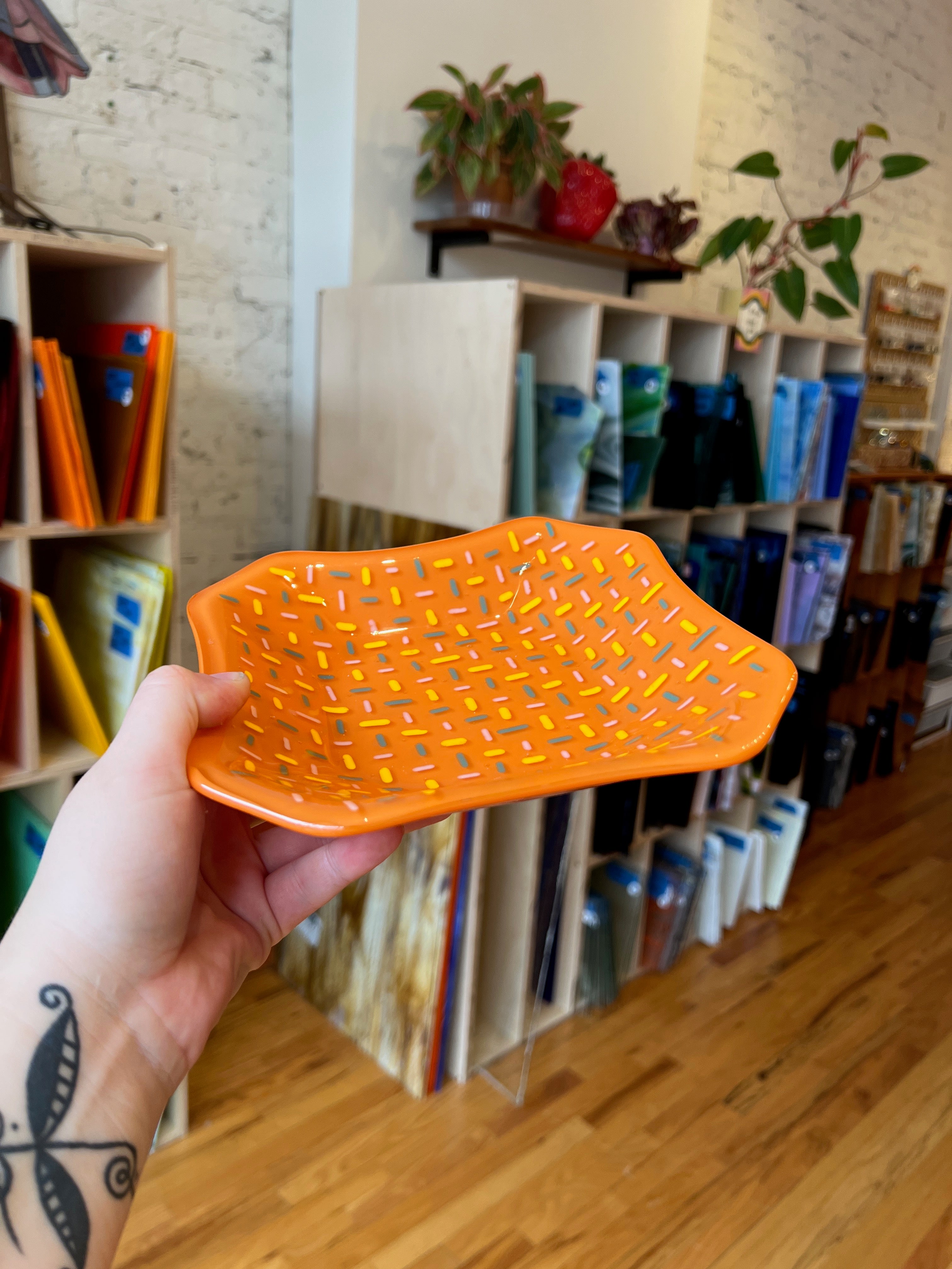 Orange fused glass tray held in a hand with a bookshelf and plants in the background