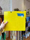 Yellow glass sheet held by a hand in a room with shelves and books.