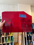 Red sheet of glass held by a hand with a price tag in a room with shelves and books.