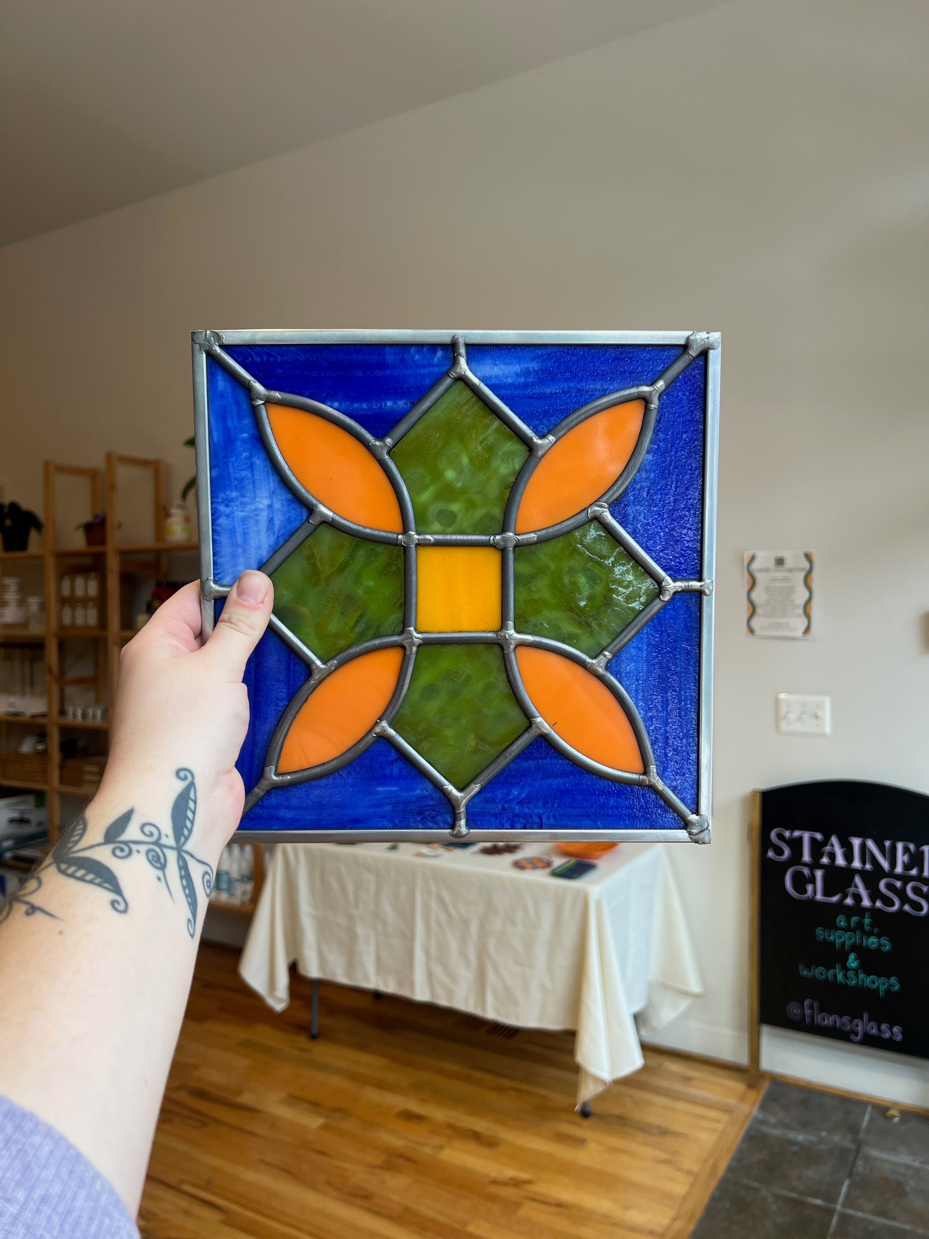 Person holding a stained glass panel with geometric design in a room with a table and sign.