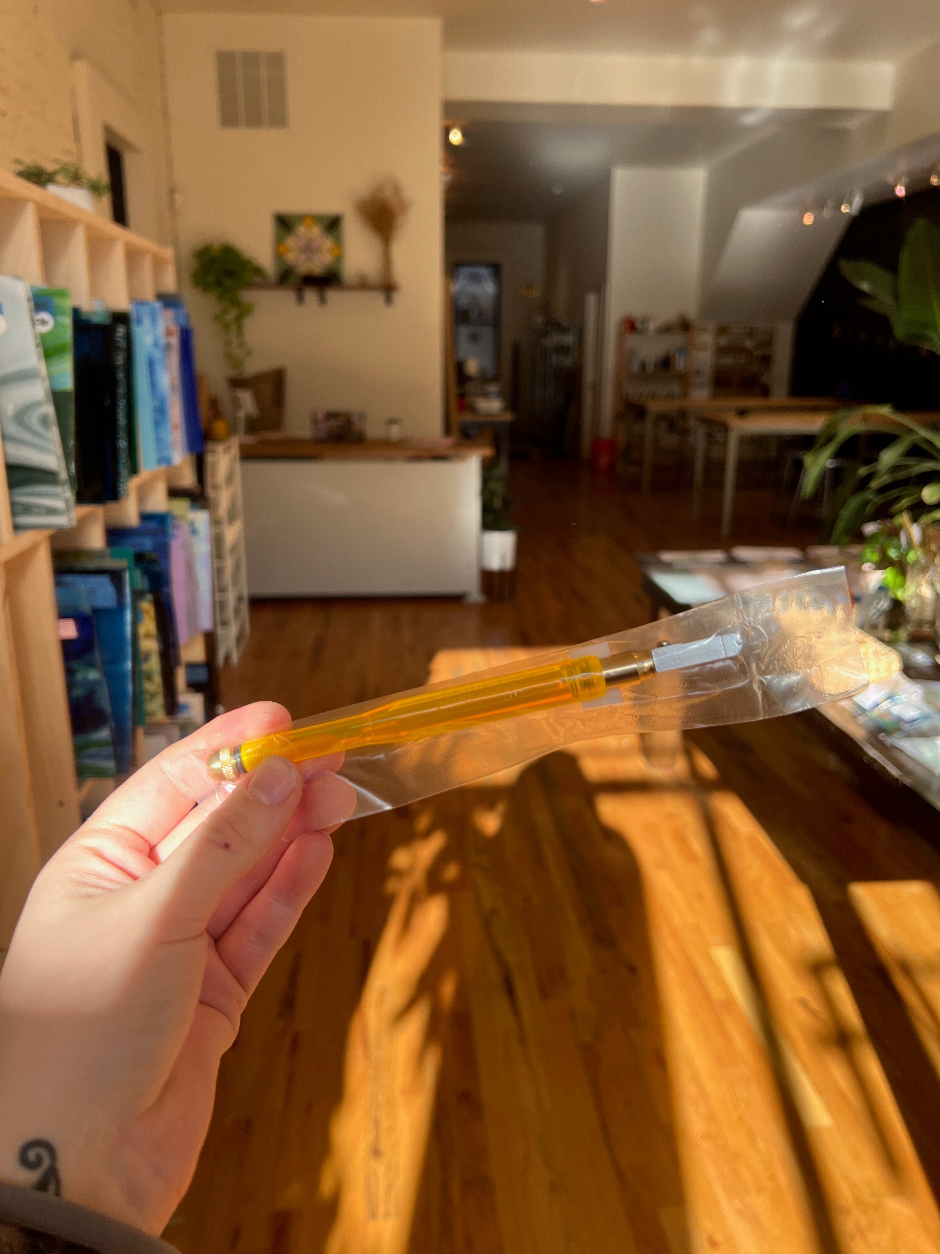 Hand holding a small yellow tube with a clear cap in a room with wooden furniture and plants.