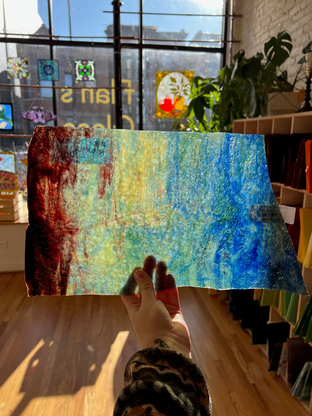 Colorful abstract sheet glass held by a person indoors with a window and plants in the background